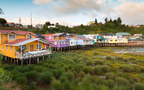 Orange, purple, blue, green, & yellow homes sit on stilts over a marsh, seen during a Patagonia & Chilean Fjords Cruise.