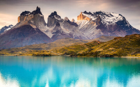 Turquoise lake with golden hillside & jagged peaks behind, seen in Torres del Paine on a Patagonia & Chilean Fjords Cruise.