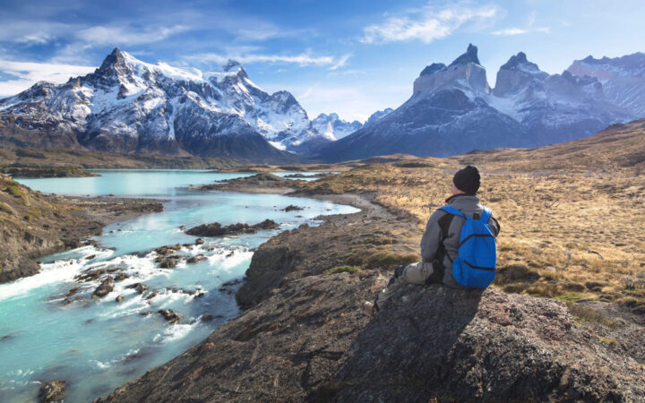 Man in gray parka, black cap & blue backpack sits on a rock by turquoise river, looking up at jagged, snowcapped mountains.