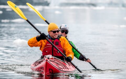 Tandem kayakers paddle in sync in a red kayak with yellow paddles & bright-colored clothes in calm water in Antarctica.