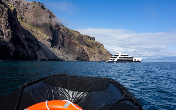 A small luxury cruise ship seen anchored next to a large cliff with a zodiac and life preserver in the foreground