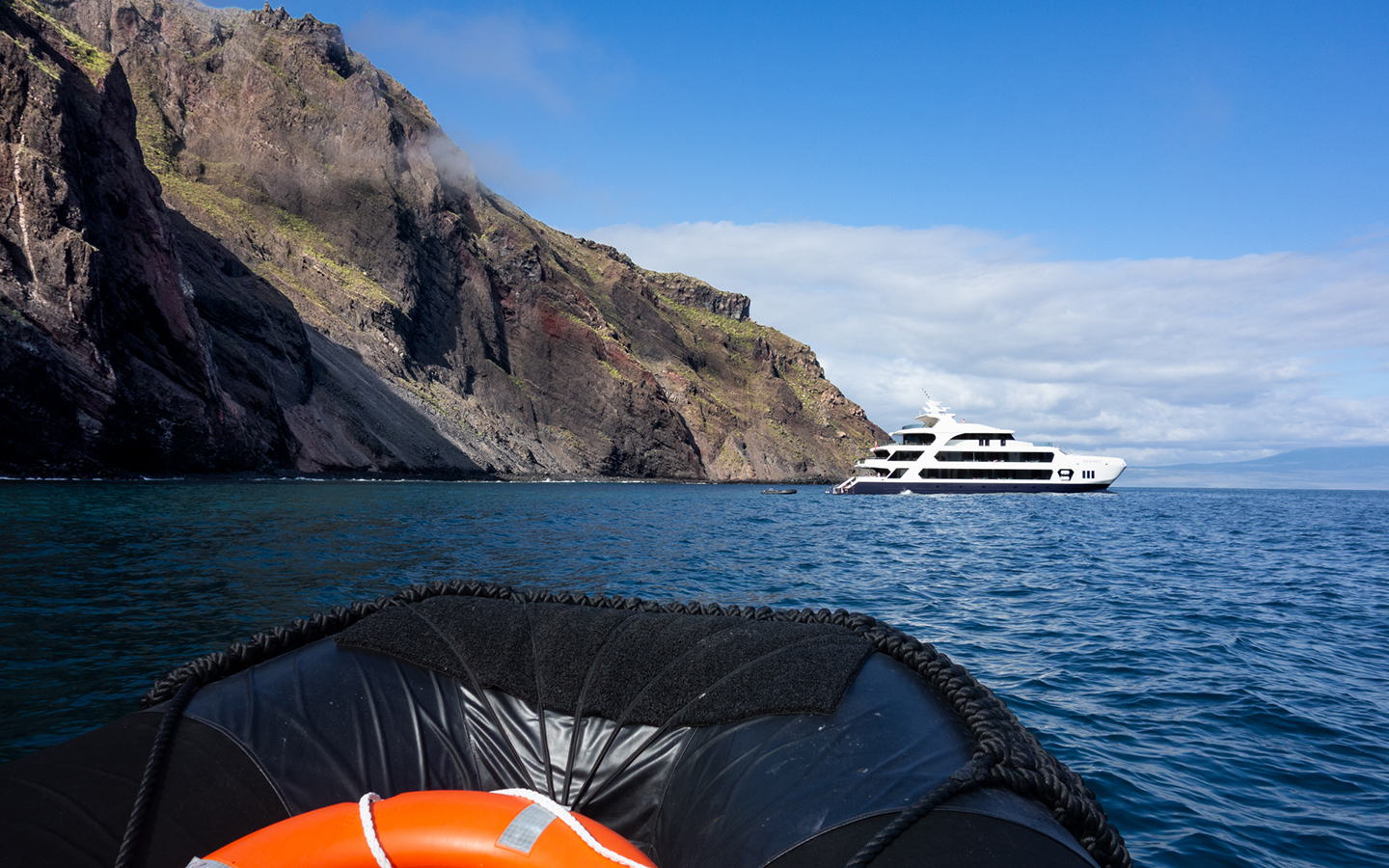 A small luxury cruise ship seen anchored next to a large cliff with a zodiac and life preserver in the foreground