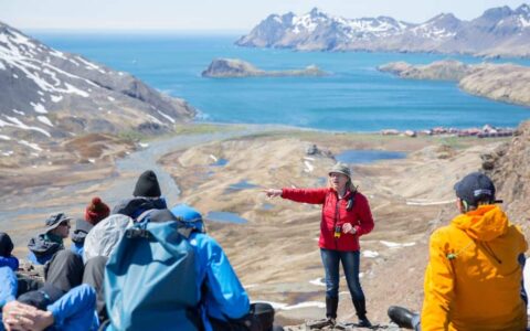 A woman in a red jacket stands & points in front of a group sitting on top of a tan mountain range during the South Georgia Antarctic Odyssey Cruise.