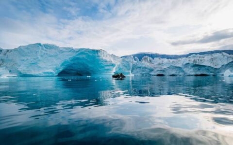 A Zodiac with polar travelers cruises past blue icebergs in a calm bay on a partly sunny day during the South Georgia Antarctic Odyssey Cruise.