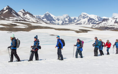 Line of In Shackleton's Footsteps cruise guests with daypacks walk over a sunny snowfield backed by jagged peaks in Antarctica.