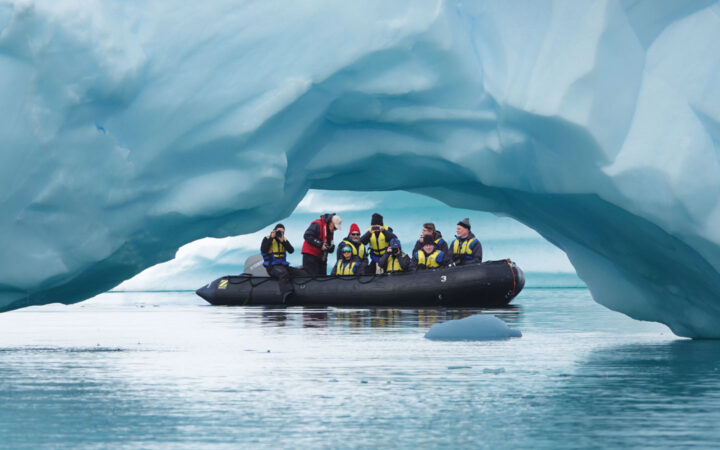 Zodiac boat with Spirit of Antarctica expedition cruise travelers dressed warmly moves past snowy arch in calm water.
