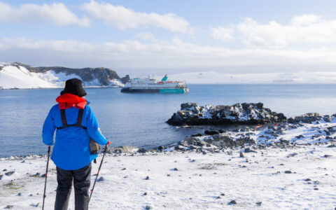 Antarctica traveler in blue jacket with red hood uses ski poles to walk over rocky, snowy ground with a small ship nearby.