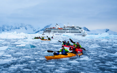 group of kayakers in brightly colored kayaks paddle through dense iceberg field. Greg Mortimer expedition ship anchored behind