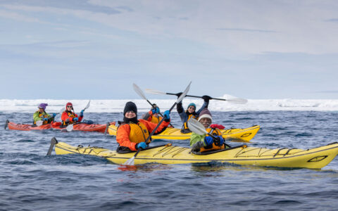 A few tandem kayakers dressed warmly paddle yellow & red boats in calm sea beside floating ice during a Svalbard cruise.