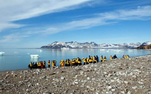 travelers stand on the arctic shore where there is a zodiac inflatable skiff waiting to take them back to the small ship