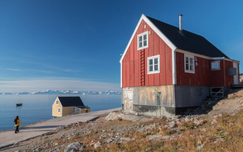3 Arctic Islands cruise guest in yellow coat walks road by water towards large red community house on a hill in the sun.