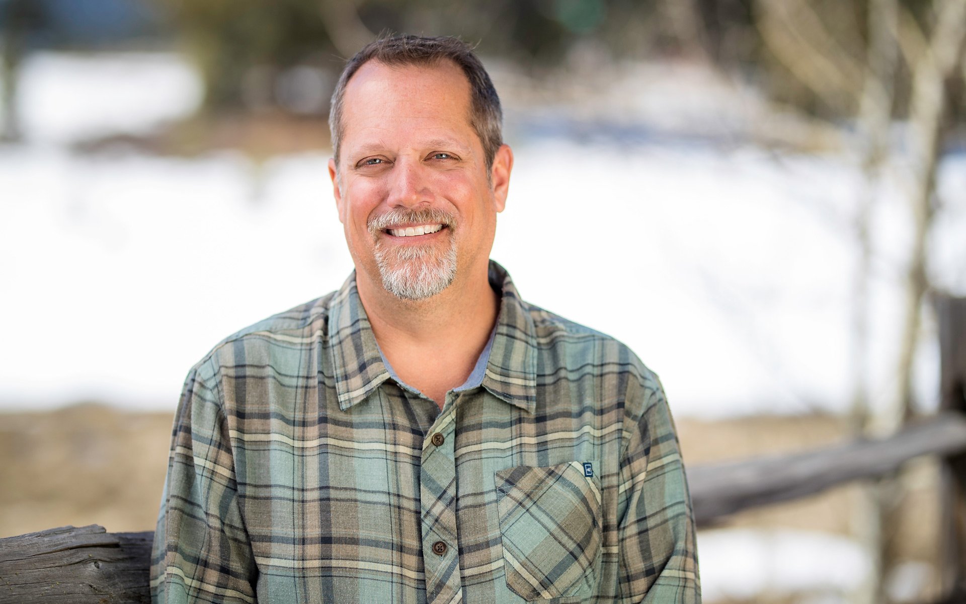 A portrait of AdventureSmith founder Todd Smith smiling looking at camera in a plaid shirt with fence behind him