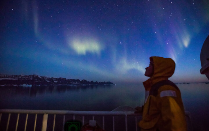 Arctic traveler in yellow polar jacket stands out on deck of ship watching northern lights of purple, blue & green in Greenland.