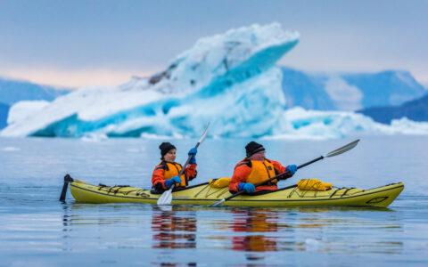 Tandem kayakers in lime green boat paddle glassy water beside a large blue iceberg during an Under The Northern Lights cruise.