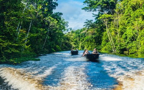 Two metal boats carry guests and cruise down the center of a river flaked on either side by lush green jungle shoreline.