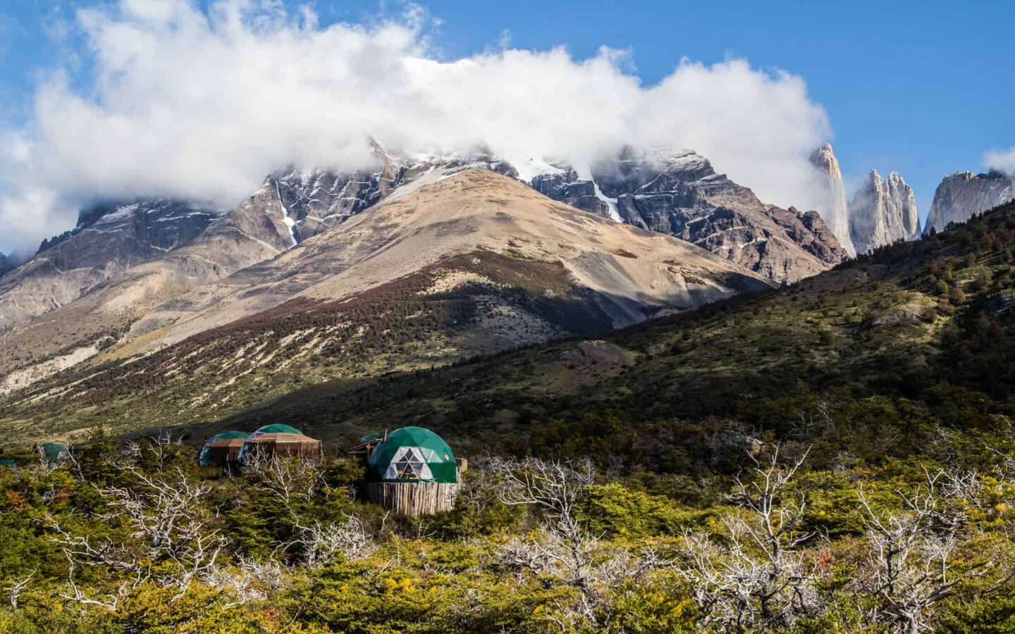 small huts that make up the patagonia ecocamp sit on a grassy hillside with large mountains in the distance