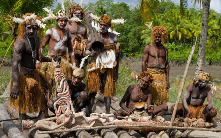Native men of the Asmat Tribe of West Papua gather around a painted wooden carving with jungle behind them on a sunny day.