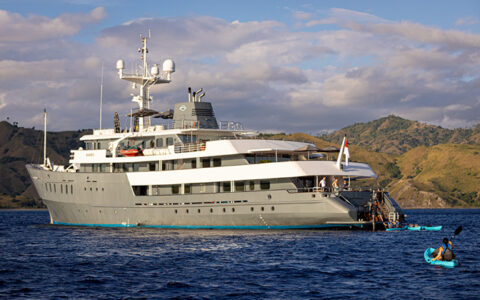 Aqua Blu small ship with gray & white exterior sits in deep blue sea offshore from beige hills as kayakers paddle nearby.