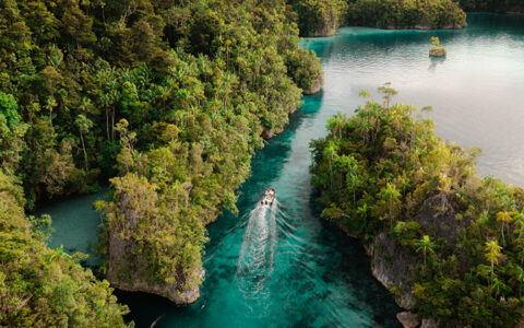Aerial view of skiff boat cruising teal sea among lush vegetation on an Aqua Blu West Papua & Asmat Tribe cruise in Indonesia.