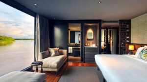 Suite on Aqua Nera Amazon luxury riverboat with king bed, 2 chaise loungers & floor-to-ceiling window looking onto river.