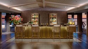 An illuminated gold bar with 4 tall bar chairs, two wall shelfs with alcohol bottles, surrounded by floor to ceiling windows.