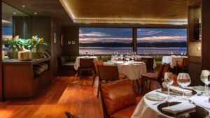 Dining room with brown leather chairs around white tablecloth covered circle tables with views outside to the purple blue sunset .