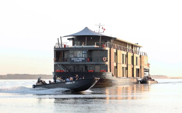 In the Peruvian Amazon, the modern three story Aqua Nera ship floats in a river as smaller metal passenger boats cruise around it.