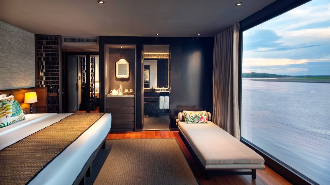 A suite aboard Aqua Nera with a panoramic floor to ceiling window with views of the river outside, king bed and small chaise lounge.