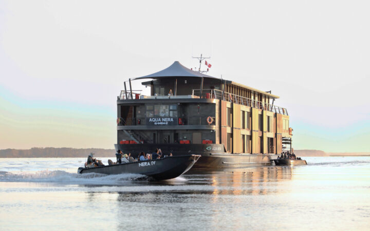In the Peruvian Amazon, the modern three story Aqua Nera ship floats in a river as smaller metal passenger boats cruise around it.