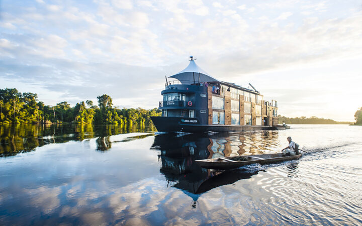 The Aria Amazon cruise ship seen anchored on still water mirroring the clouds in the sky with a local guide in a skiff in front of the ship.