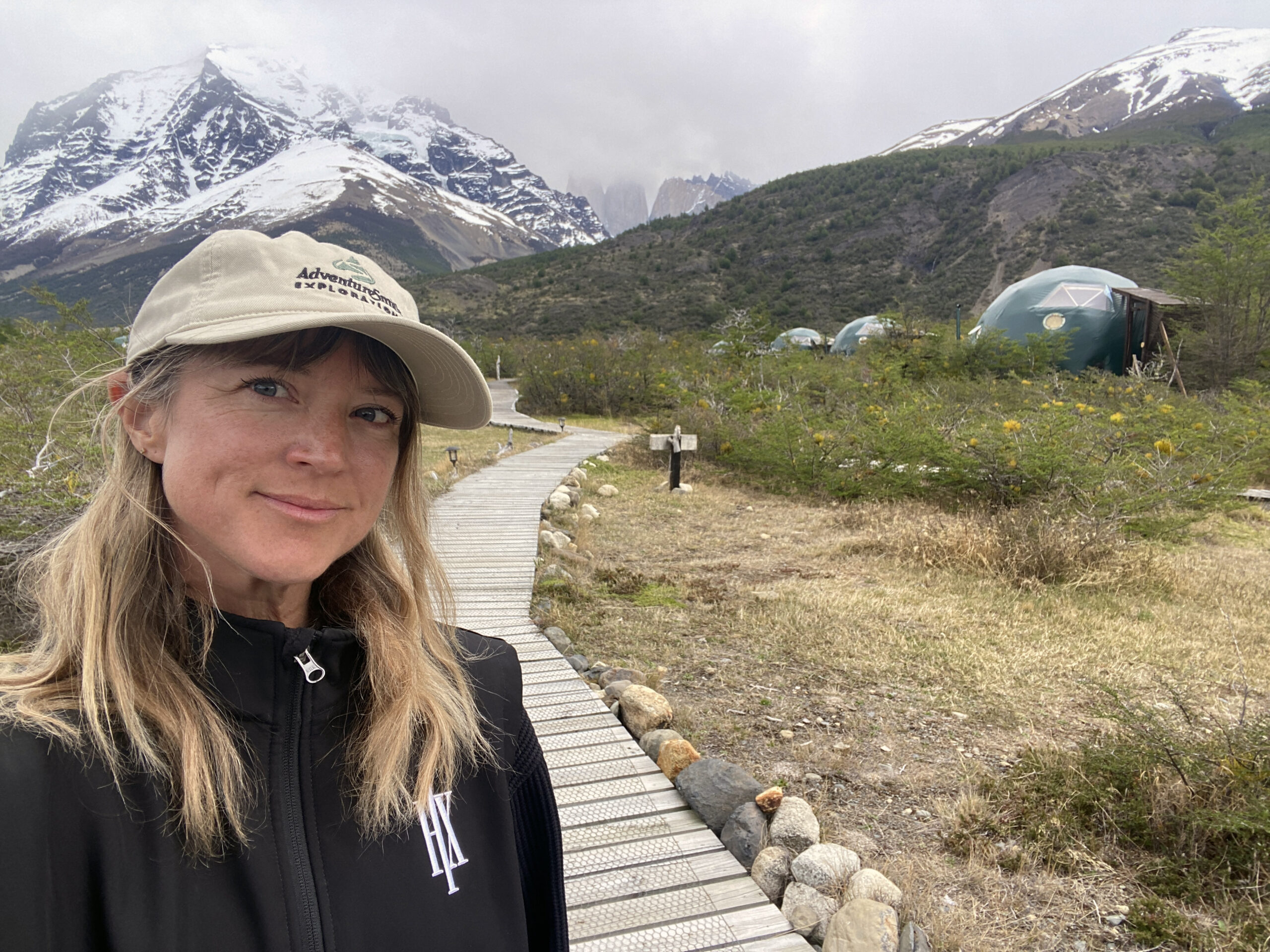 A female traveler in a tan AdventureSmith hat with EcoCamp domes and boardwalk seen behind her with a cloudy sky