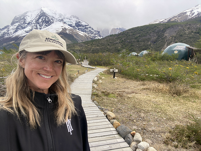 A woman in a tan AdventureSmith hat and black jacket stands on boardwalk path at EcoCamp Patagonia with green domes behind her