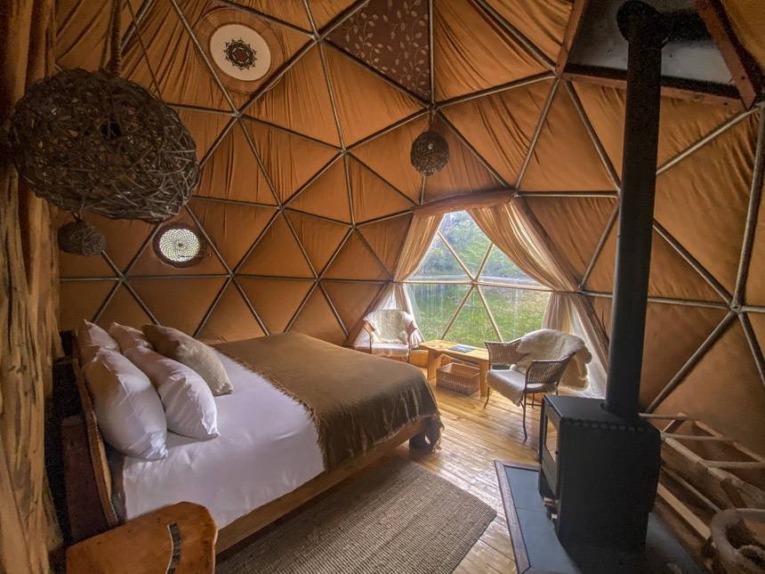 Inside of the dome EcoCamp with a bed, chairs, table and wood burning stove looking out into the forest of Torres del Paine.