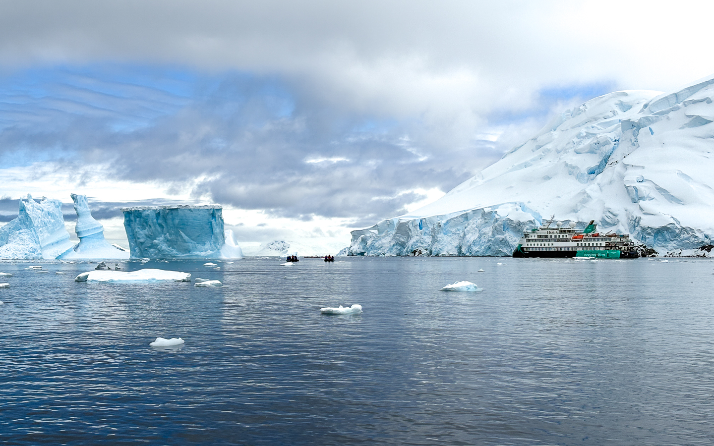 Sylvia Earle off in the distance in Antarctica with snow covered and giant icebergs and mountains on a cloudy calm day