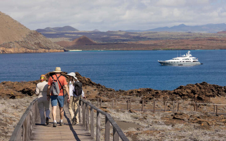 Aqua Mare Galapagos cruise travelers in shorts & t-shirts walk a wooden boardwalk over arid land as yacht sits offshore.
