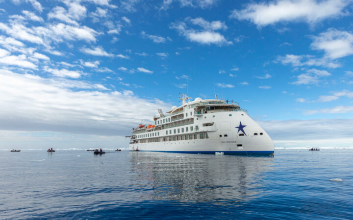Greg Mortimer ship white and blue exterior with zodiacs floating by on a calm and cloudy day in antarctica