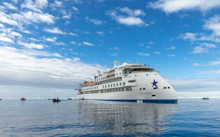 Greg Mortimer ship white and blue exterior with zodiacs floating by on a calm and cloudy day in antarctica