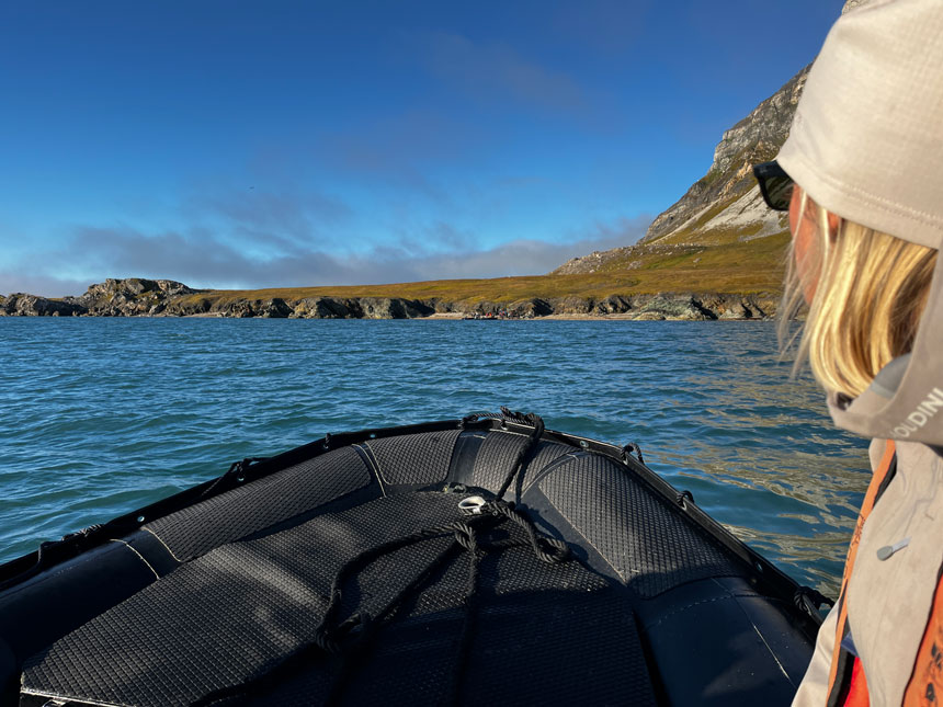 Blonde woman in white outerwear rides Zodiac boat towards green shoreline on a Svalbard cruise.