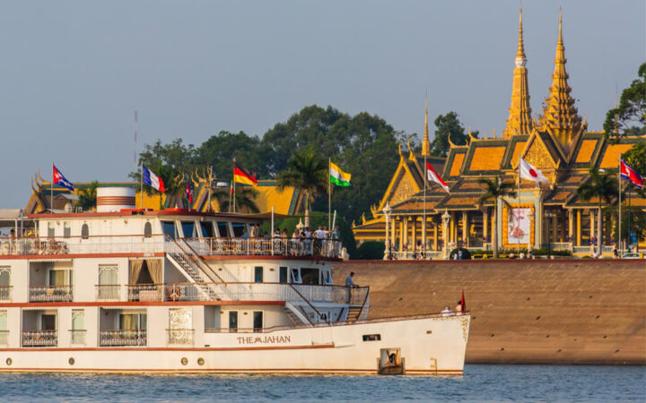 The Jahan river boat cruises the Mekong river in Cambodia with guests on the bow, an intricate golden temple is seen onshore.