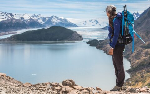 Young woman wearing a day pack with trekking poles stands on a rocky summit looking down on a lake on the Torres del Paine Trek.