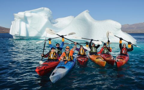 A group of kayakers in red orange and blue kayaks hold their paddles in the air as they float in front of a large iceberg in Antarctica