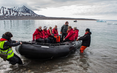 Guides stand in shallow water & pull black Zodiac boat with Arctic Highlights cruise guests in to shore in cloudy Svalbard.
