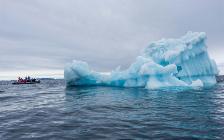 An iceberg rises out of a grey ocean next to an inflatable boat full of passengers from the expedition around spitsbergen