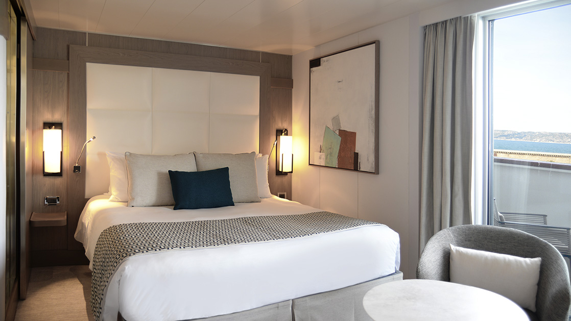 Deluxe stateroom aboard L'Austral expedition ship, showing king bed, table, chair & white-&-copper appointments.