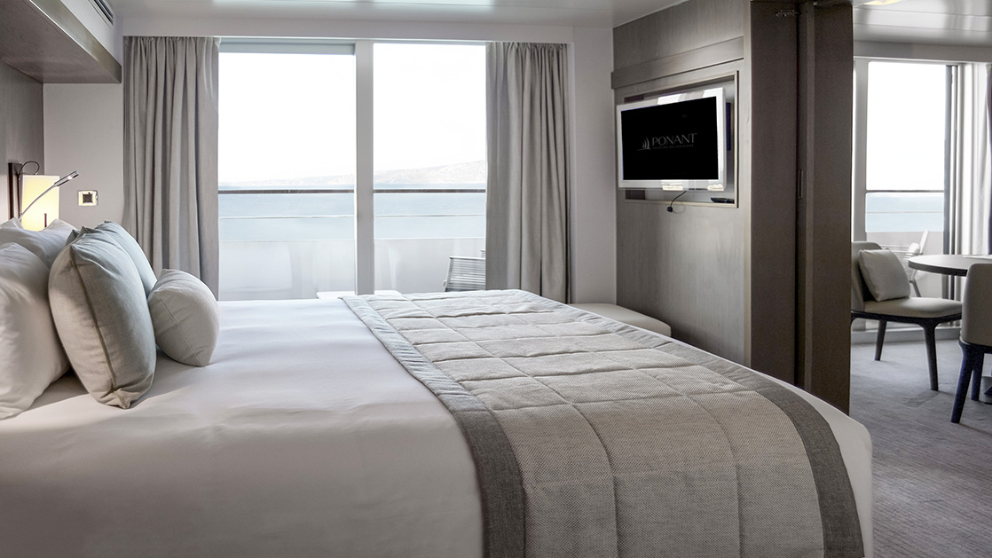 Deluxe Suite aboard L'Austral expedition ship, showing king bed, table, TV, chair & white-&-copper appointments.