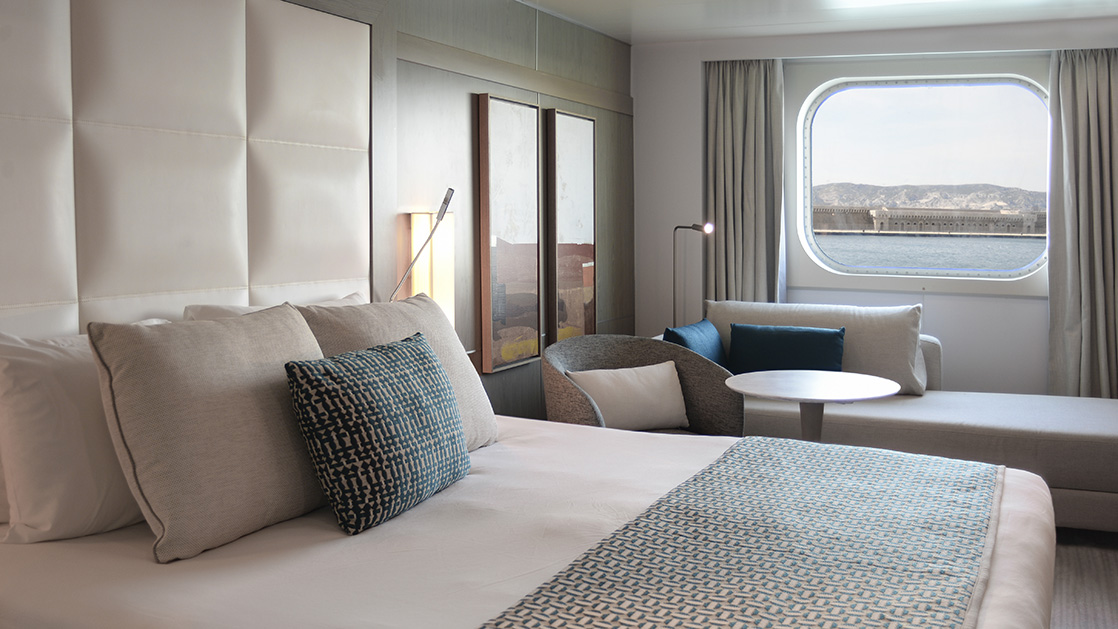 Superior stateroom aboard L'Austral expedition ship, showing king bed, desk, lamp & white-&-copper appointments.