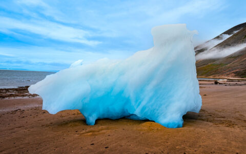 Bright blue iceberg bit washed up on brown shoreline under a blue sky in Baffin Bay, Greenland.