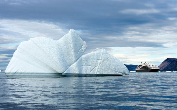 Cruising in Greenland's Baffin Bay is a luxury Ponant ship, dwarfed in size compared to a large iceberg floating next to it.