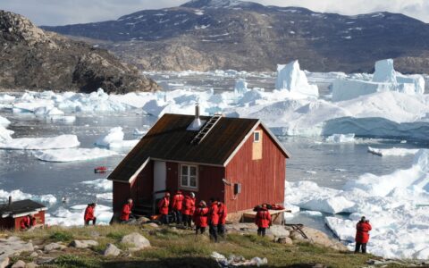Luxury ponant cruise guests in red parkas explore the shore in Greenland among red wood building and a sea filled with icebergs.