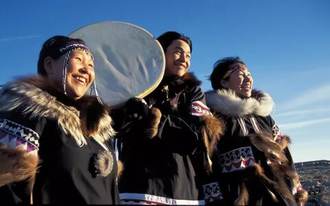 Three Inuit women, arctic indigenous people of Greenland, dressed in traditional clothing and hold a musical instrument.